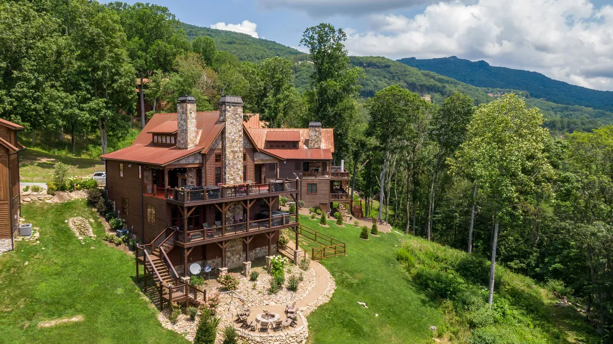The image showcases a large, inviting wooden house with a striking stone chimney nestled among mature trees. The spacious deck features comfortable outdoor seating, perfect for gathering with family or friends. Surrounded by nature, the home exudes a relaxed, communal atmosphere ideal for enjoying sunset views, early morning coffee, or evenings under the stars.