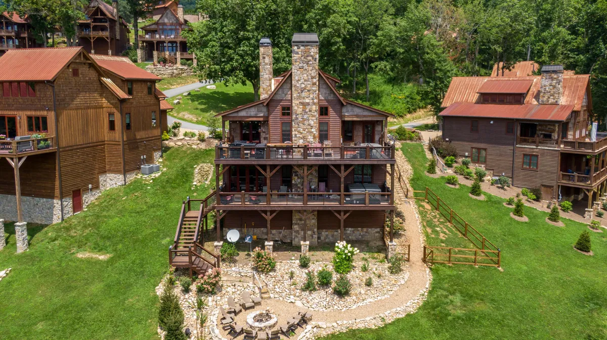 A large stone chimney sets the rustic tone for this spacious deck and fire pit area, enveloped by lush greenery. The image captures multiple seating options, ideal for group barbecues, outdoor games, or simply gathering around the fire pit to extend the fun into the night.