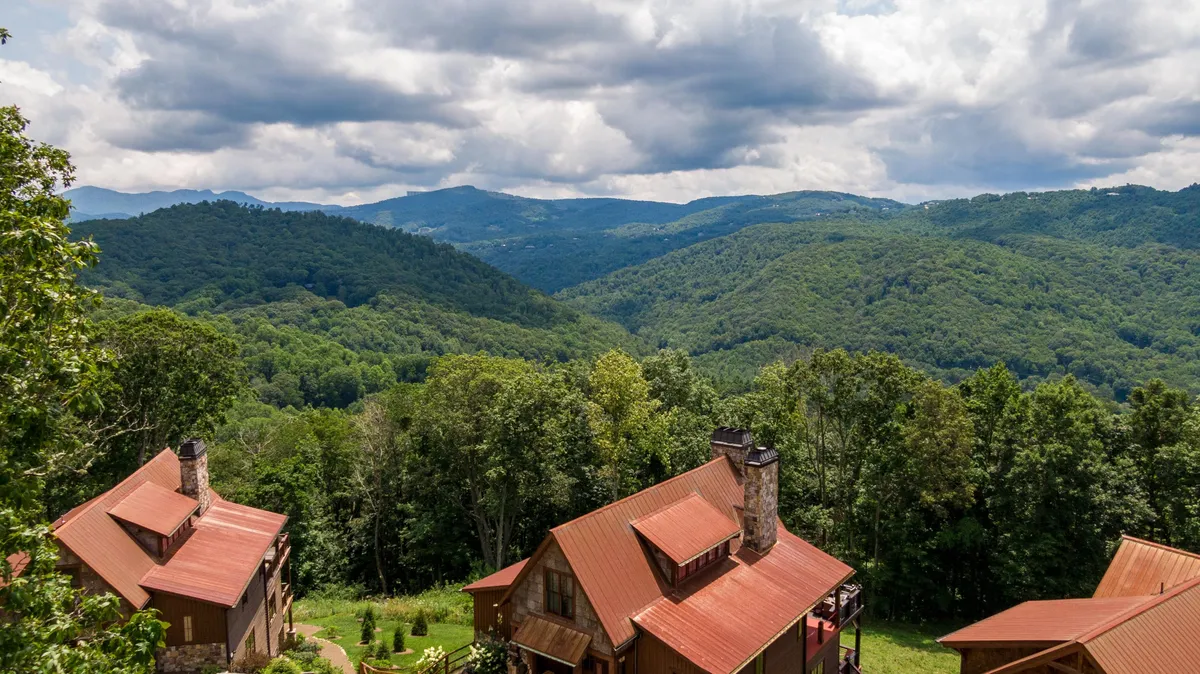 A wide exterior shot featuring rustic cabins nestled among rolling green hills, with distant mountains and a blue sky dotted with clouds. The natural landscape is lush and inviting, evoking a sense of peaceful seclusion and expansive outdoor adventure.