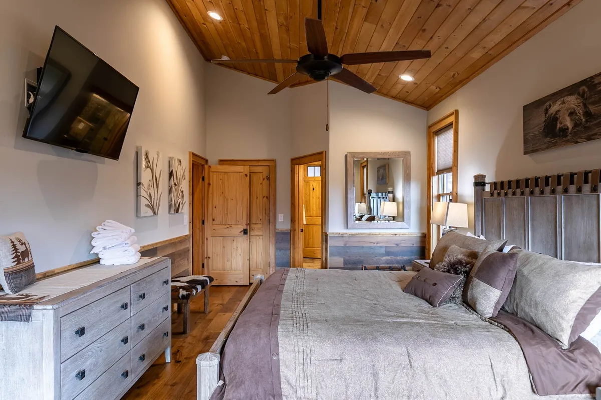 A tranquil bedroom featuring a king-sized bed with plush bedding, a wooden ceiling, a large wall-mounted TV, a decorative mirror, and wooden dressers and doors. The atmosphere is serene and inviting, ideal for restful nights and cozy mornings.