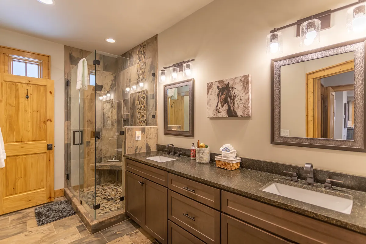A modern bathroom with dual vanities, a walk-in shower, tasteful wall art, and a wooden door, offering both functionality and a serene environment for guests to relax and refresh.