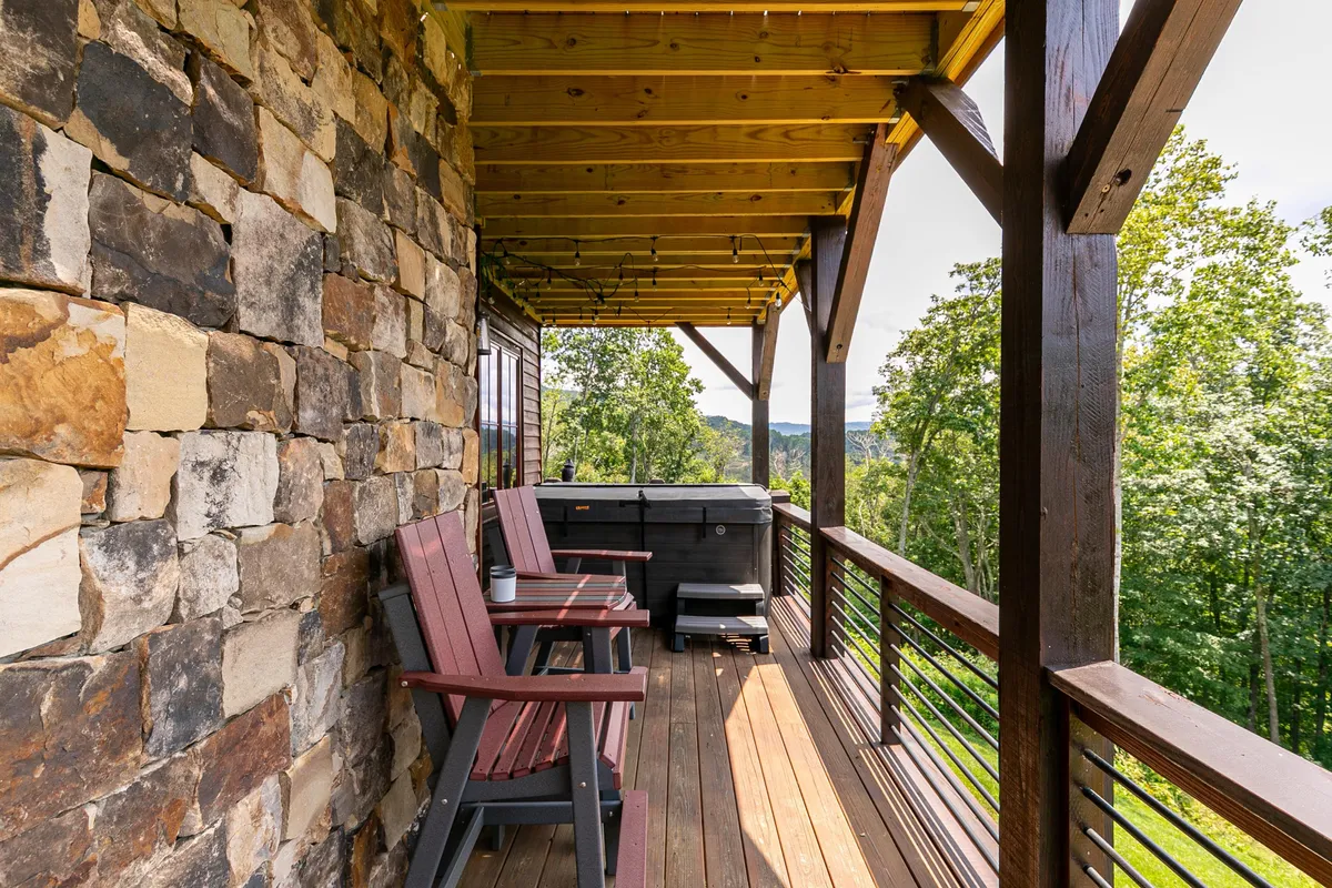 An exterior scene of a wooden deck with a hot tub, stone wall, and adirondack chairs, opening onto serene natural views. This space is set up for relaxing outdoors, soaking in the hot tub, and enjoying tranquil moments close to nature.
