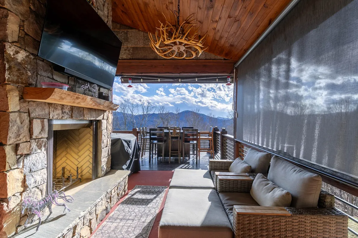 An inviting main-level deck featuring a stone fireplace, comfortable seating, a dining table, and expansive mountain views. This outdoor living area is ideal for alfresco dining, relaxing by the fire, and soaking in the surrounding scenery.