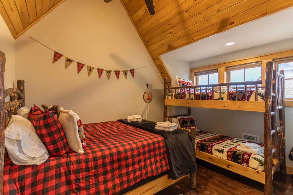 A playful bunk room with a twin-over-twin bunk bed, plaid bedding, sturdy wooden beams, and cheerful decorative bunting. The space feels inviting and fun, ideal for kids, teens, or extra guests.
