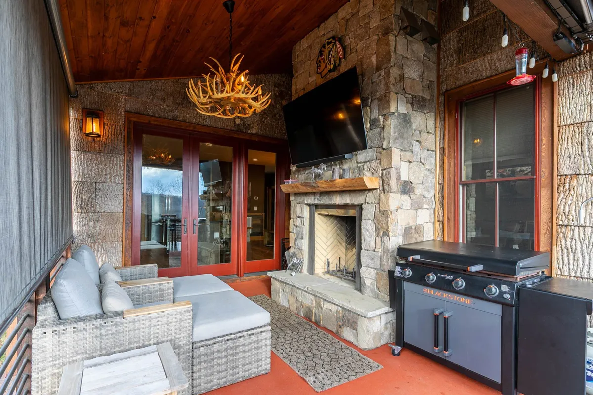 An inviting outdoor seating area centers around a stone fireplace. Adirondack chairs and a grill suggest the perfect spot for evening gatherings, outdoor dining, or simply relaxing by the fire under the stars. Lush surroundings and thoughtful lighting enhance the communal and cozy feel of the space.