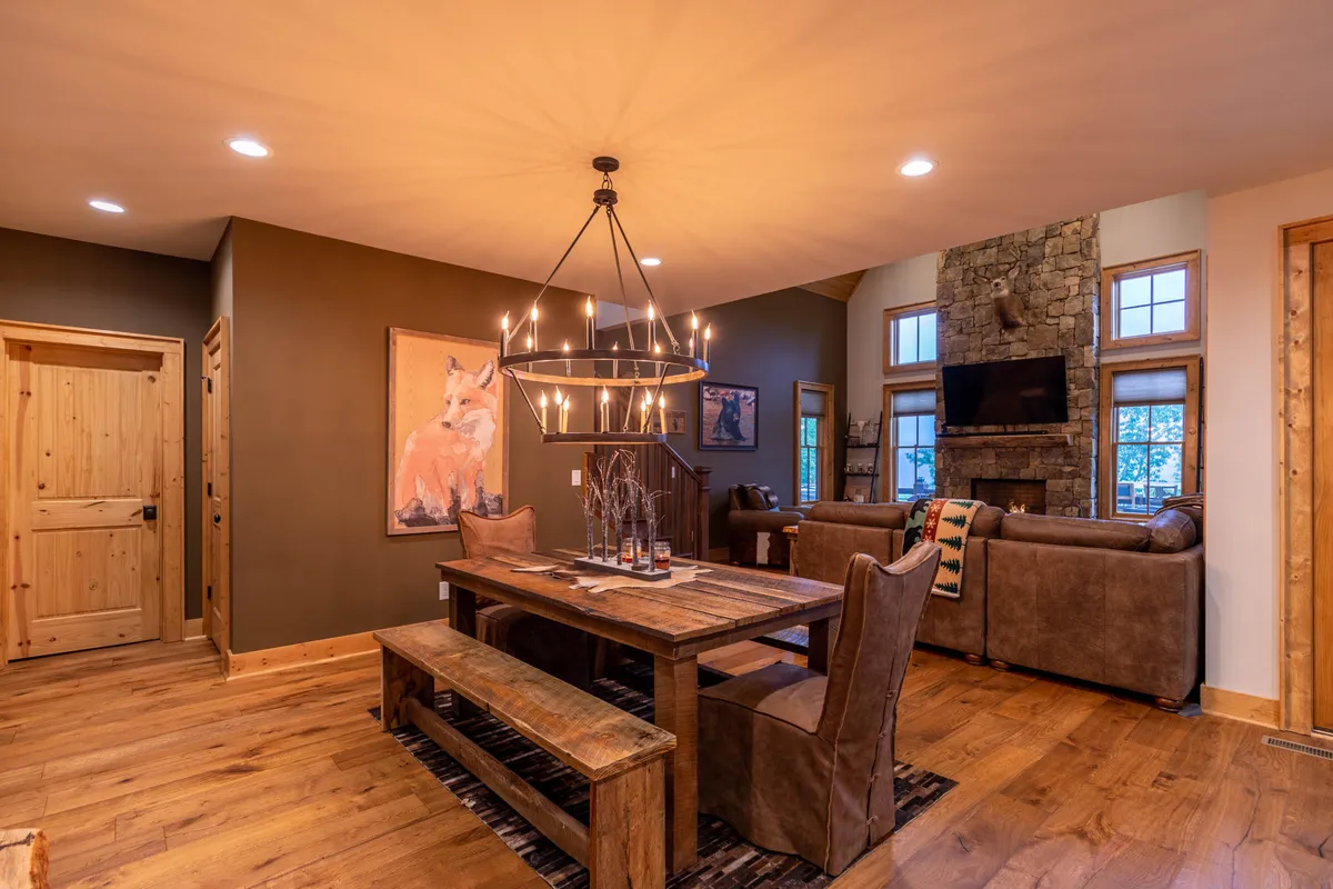 A large wooden dining table under a glowing chandelier, flanked by a striking stone fireplace and warm rustic decor. The arrangement emphasizes togetherness, with enough space for 6–8 people to dine, set in a cozy, inviting atmosphere.