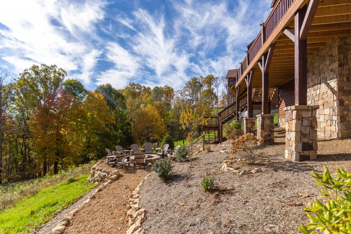 This image showcases a spacious stone patio and wooden deck surrounded by lush trees, complete with Adirondack chairs for group seating. The setting is perfect for evening gatherings, watching the sunset, and socializing around a fire pit, with a backdrop of natural beauty.