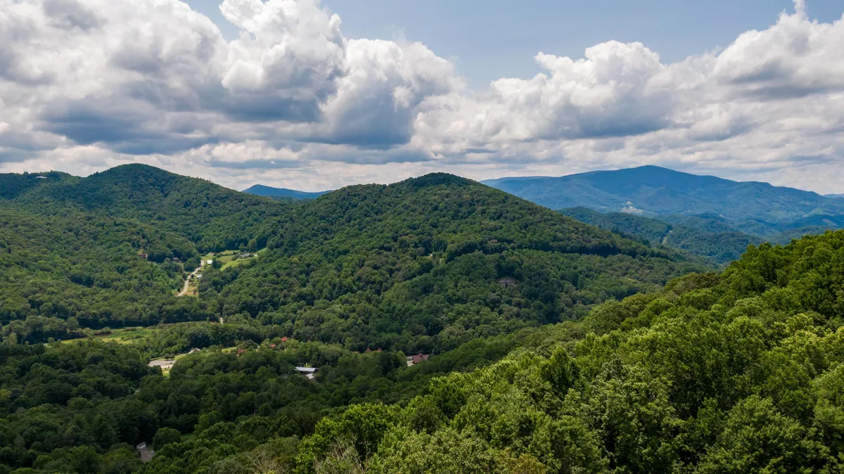A scenic view of rolling green hills and distant mountains under a cloudy sky. The landscape is picturesque, highlighting the natural splendor of the area and showcasing seasonal color changes.